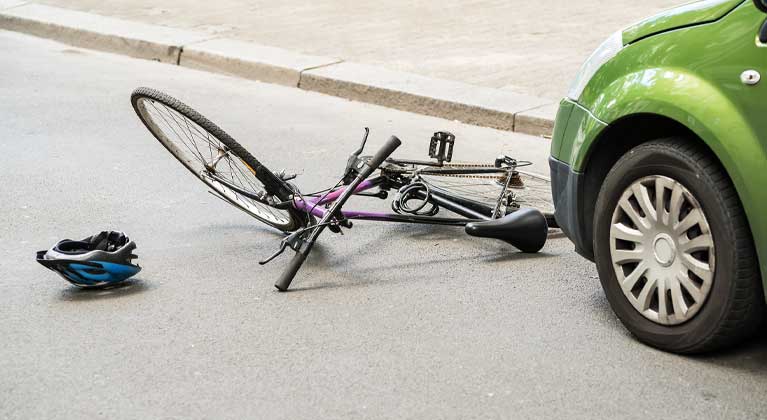 Bicicleta morada tirada en la calle junto a la llanta de un carro verde, con un casco azul en el pavimento después de un choque