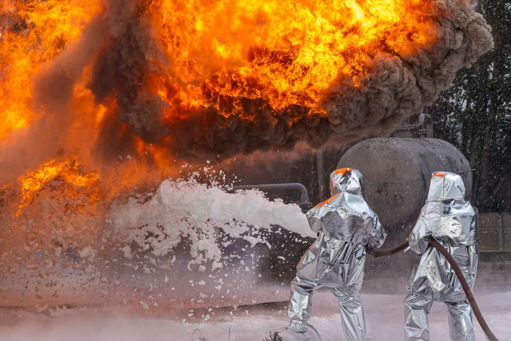 Two firefighters in silver heat-protective suits spraying foam on a large fire next to an industrial tank