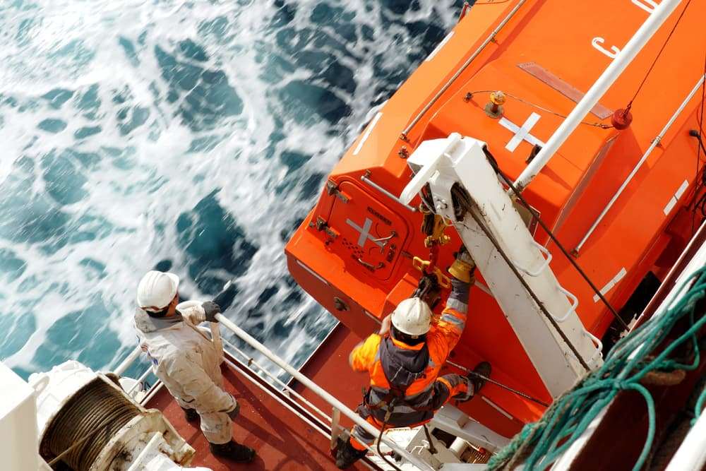 Crew member with orange jacket and helmet checking aft hook on life boat fixed to container vessel