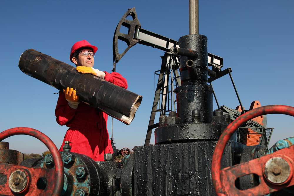 Trabajador de campo petrolero con overol rojo cargando un tubo junto a una bomba de extracción