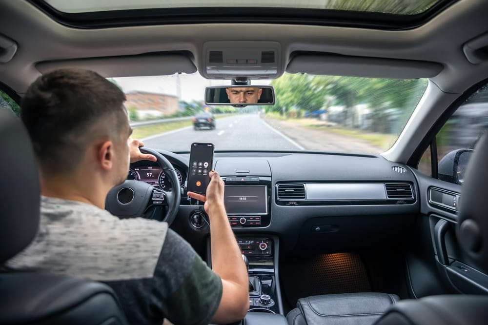 Hombre manejando en carretera mientras mira el celular que sostiene junto al volante
