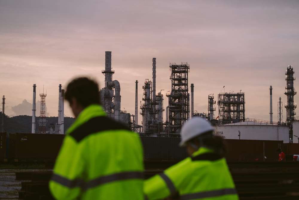 Two workers in high-visibility jackets and a hard hat looking toward refinery towers at dusk