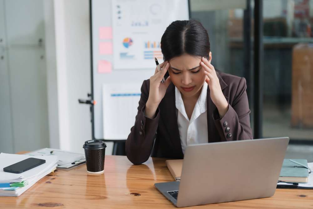Office worker holding her head while experiencing a headache or stress at her desk in the workplace.