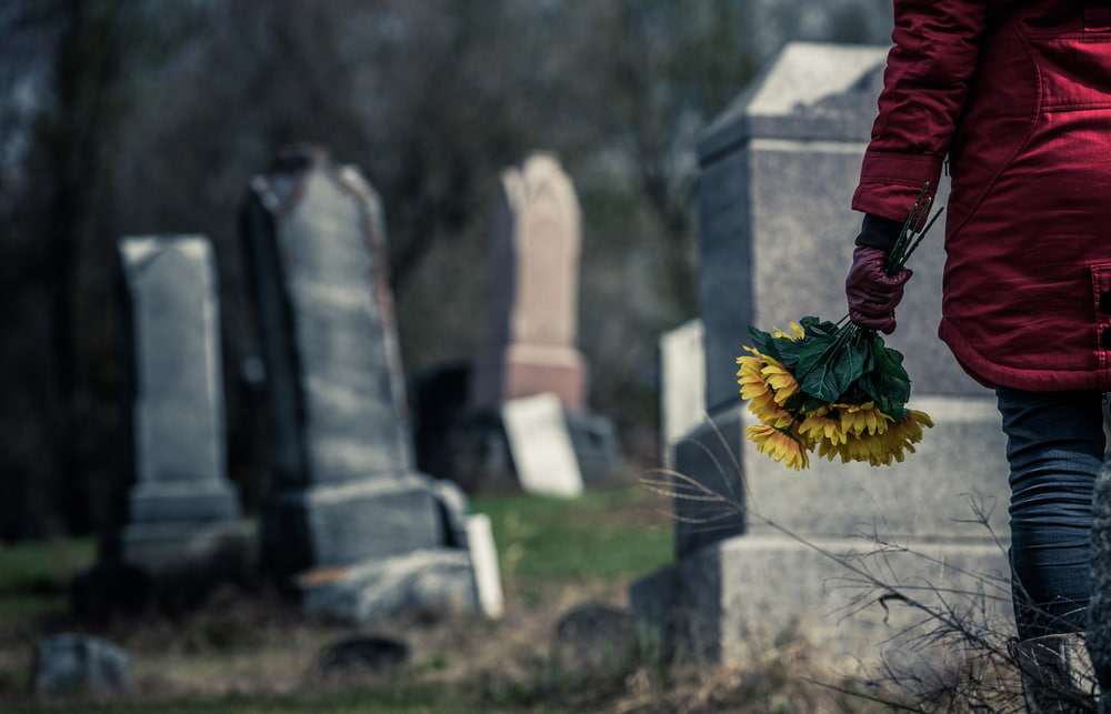 Person in a red coat carrying a bouquet of sunflowers while visiting a grave in a cemetery