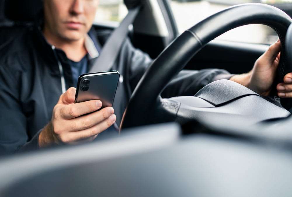 Conductor con chaqueta oscura revisando su celular con una mano mientras la otra sostiene el volante