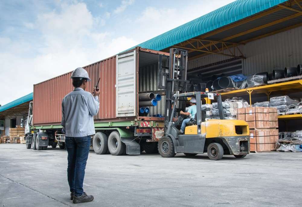Forklift loading materials into a shipping container while a worker supervises the operation