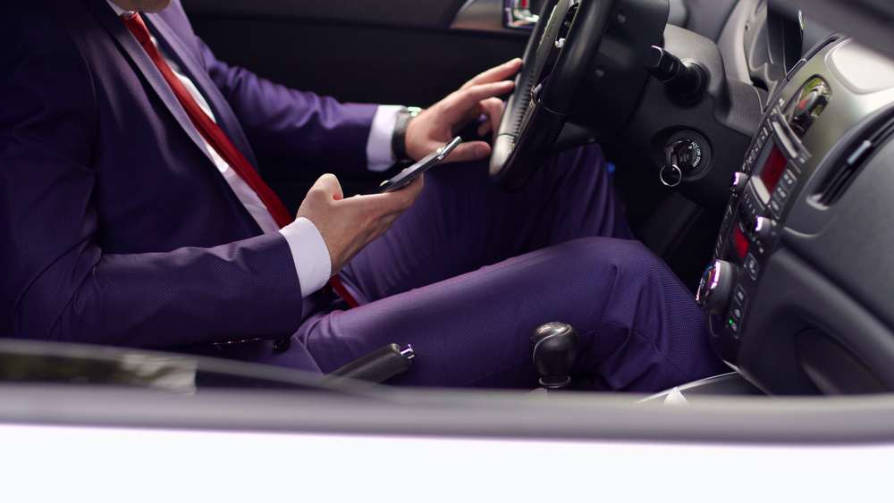 Hombre de traje morado y corbata roja usando su celular con una mano mientras sostiene el volante